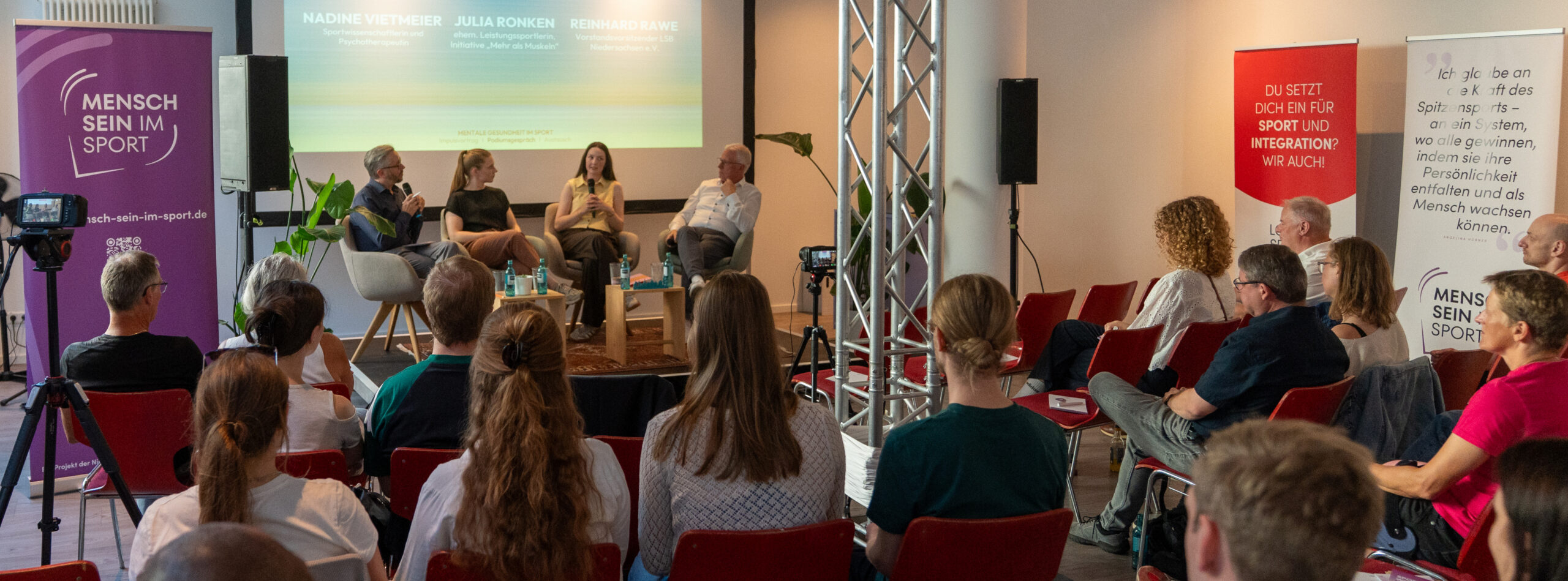Publikum sitzt in einem Raum und hört einem Podiumsgespräch mit vier Personen auf einer Bühne zu