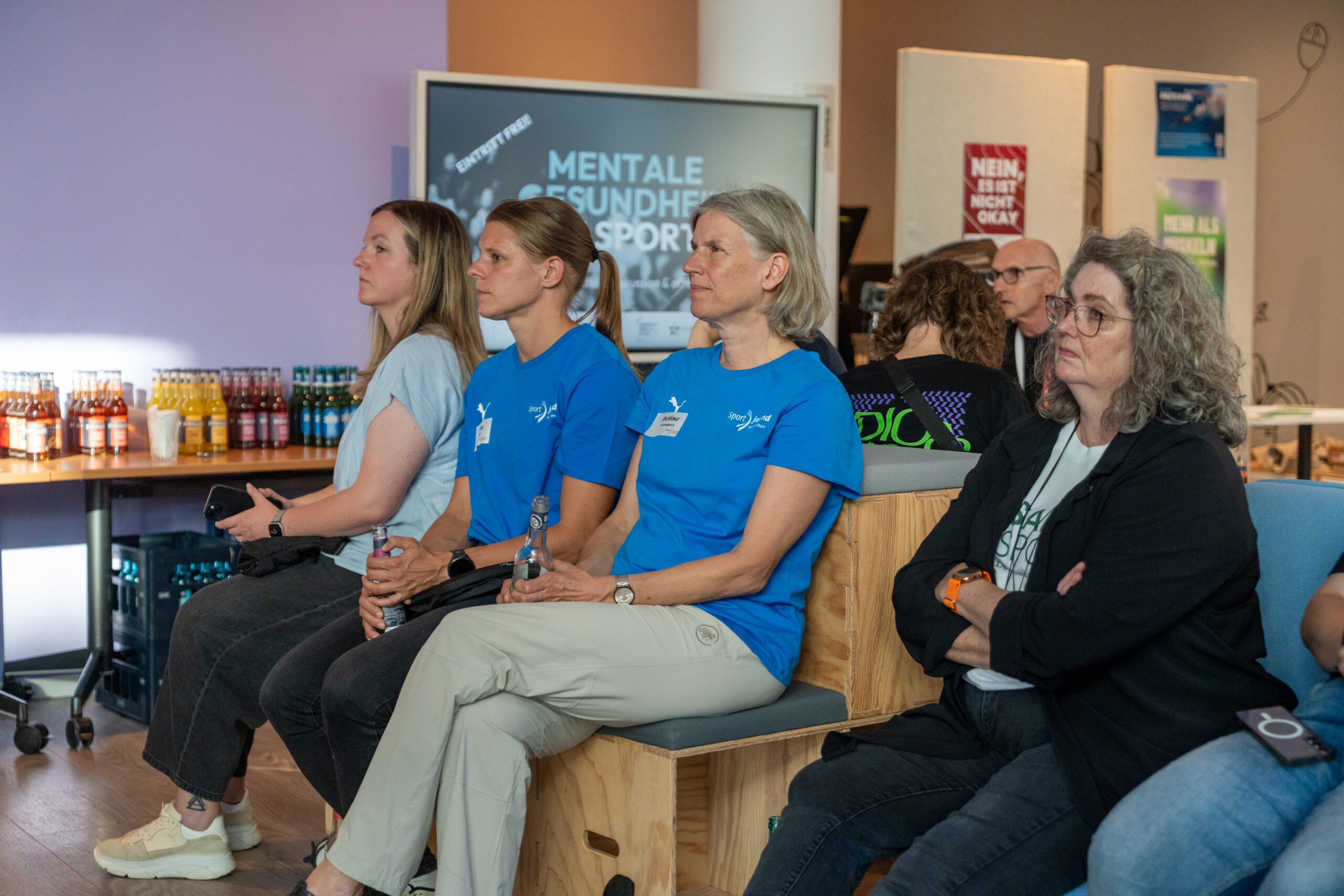 Drei Frauen sitzen nebeneinander auf einer Bank, zwei tragen blaue T-Shirts mit Namensschildern und halten Wasserflaschen, im Hintergrund ein Bildschirm mit der Aufschrift 'MENTALE GESUNDHEIT SPORT'