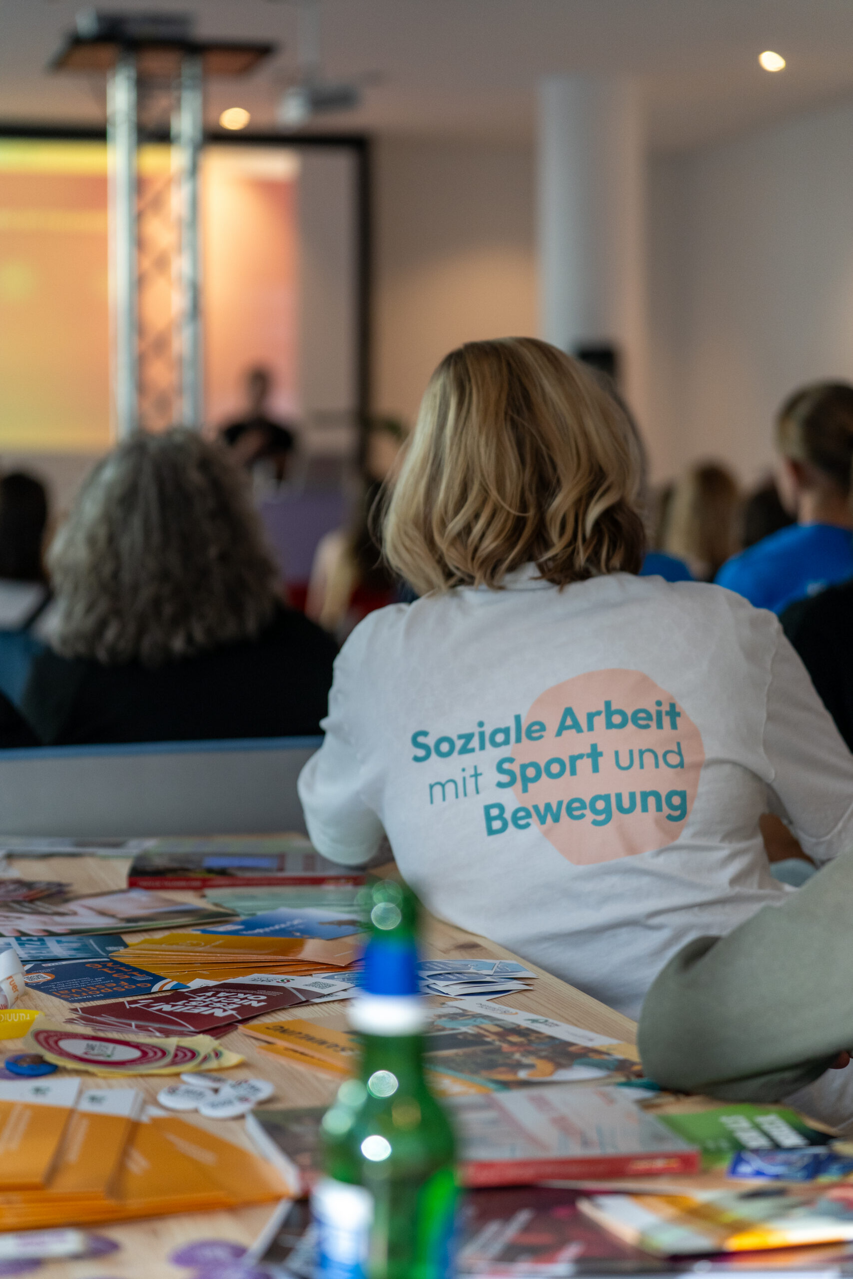 Person mit hellblondem schulterlangem Haar sitzt in einem Publikum und trägt ein weißes T-Shirt mit dem Schriftzug 'Soziale Arbeit mit Sport und Bewegung'.